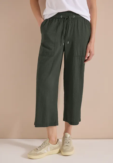 Unterschenkel einer Person in lässiger, knöchellanger, olivgrüner Hose mit Kordelzug, dazu cremefarbene Sneaker.