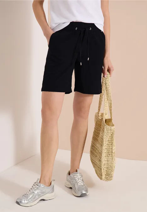 Person in schwarzen Shorts und weißem T-Shirt, silbernen Sneakern, hält geflochtene Strohtasche vor hellem Hintergrund.