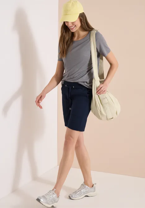 Junge Frau in grauem T-Shirt, dunkelblauen Bermudas, Sneakern, heller Tasche und Cap vor neutralem Hintergrund.