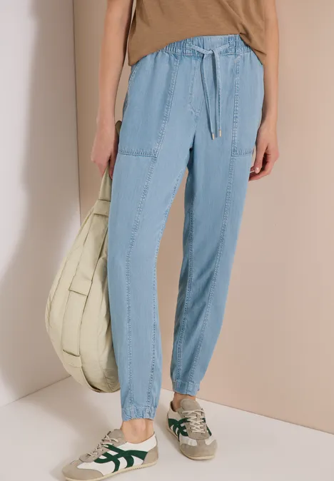 Persoon in lichtblauwe cargobroek met boordjes, beige top, groene sneakers en grote stoffen tas.