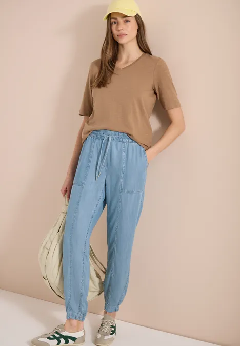 Vrouw in beige T-shirt, blauwe joggingbroek en sneakers, met een grote tas.