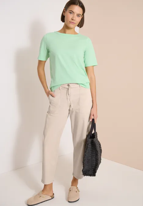 Person trägt mintgrünes Shirt, beige Hose und Schuhe, hält schwarze Handtasche vor hellem Hintergrund.