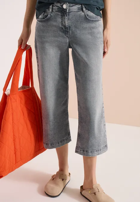 Weite graue 7/8-Jeans, orangene Tasche in der Hand und beige Hausschuhe auf hellem Hintergrund.