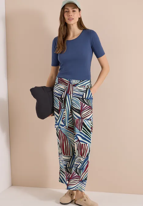 Femme en haut bleu, pantalon ample à motifs et espadrilles, tenant un chapeau bleu marine.