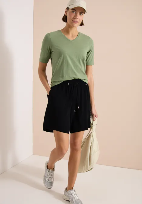 Personne portant un t-shirt vert en V, short noir, baskets argentées et un sac en toile sur fond neutre.