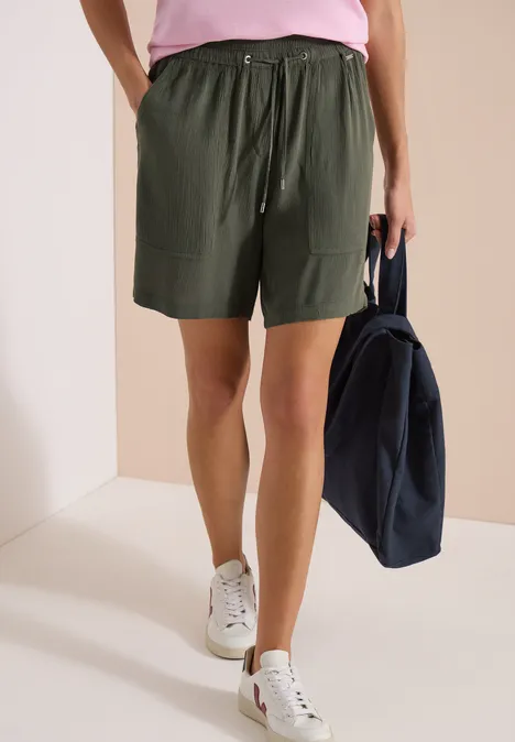 Vrouw in groene short, roze T-shirt en witte sneakers, met een donkerblauwe tas.