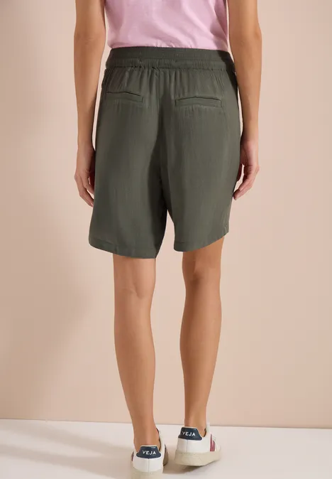 Achteraanzicht van persoon met losse kaki short, roze top en witte sneakers voor een beige achtergrond.
