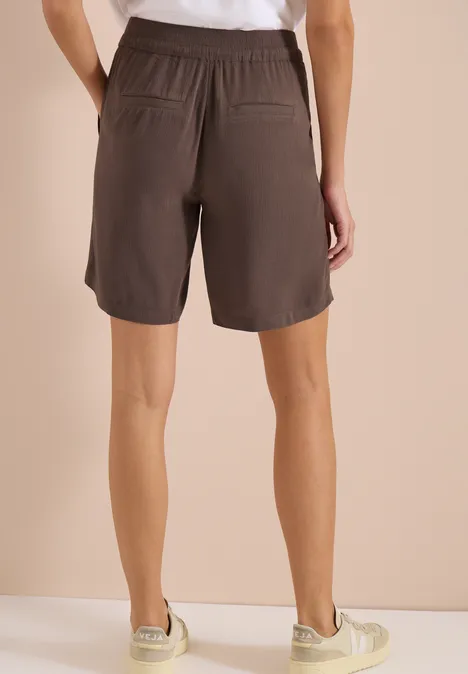 Rückenansicht einer Person in lässigen braunen Shorts mit elastischem Bund und Taschen vor beigem Hintergrund.