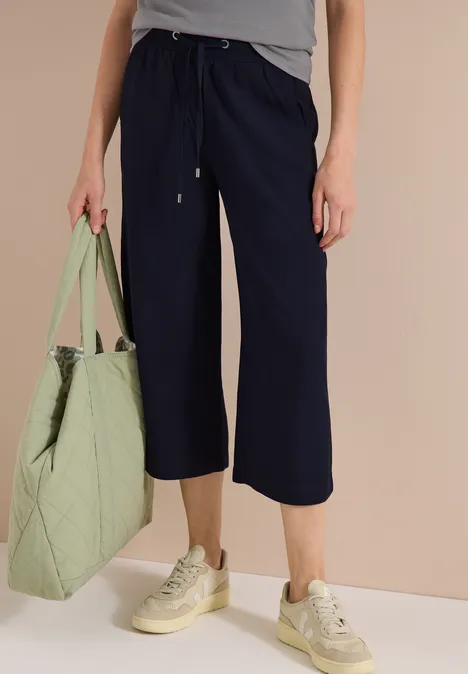 Frau trägt dunkelblaue Culotte-Hose, graues Top, beige Sneaker und hält eine große grüne Tasche.