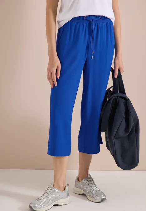 Persoon draagt blauwe 7/8-sweatpants, wit shirt, grijze sneakers en heeft een blauwe sporttas bij zich.