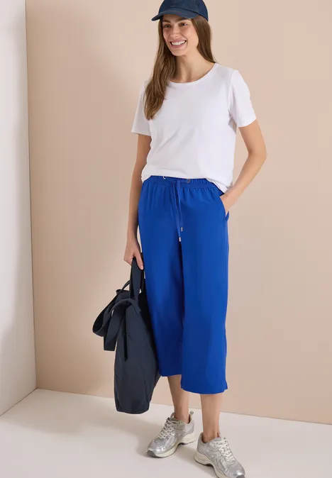 Vrouw in wit T-shirt en blauwe wijde broek, sneakers, draagt tas voor een beige-witte achtergrond.