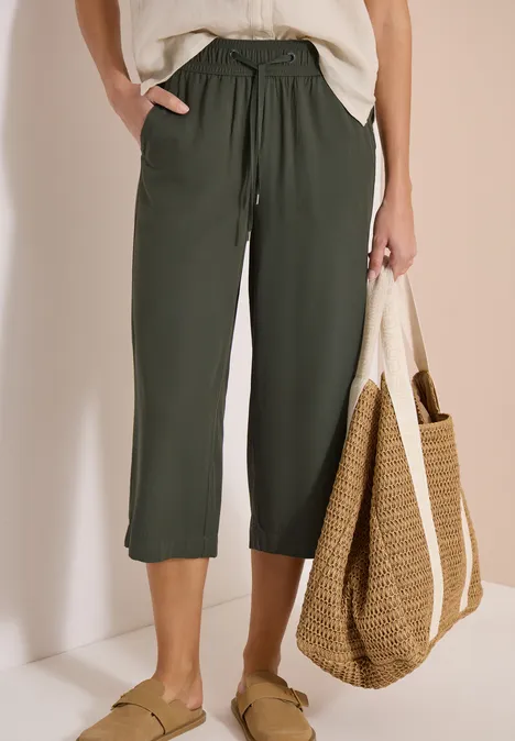 Femme en pantacourt kaki à cordon, top blanc, grand sac en paille et mules vertes