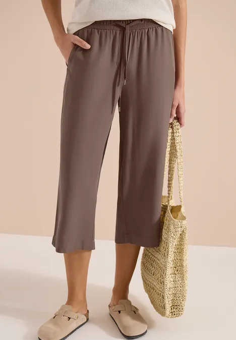 Bruine, wijde 3/4-broek met elastische taille, gecombineerd met beige instappers en een gevlochten strozak.