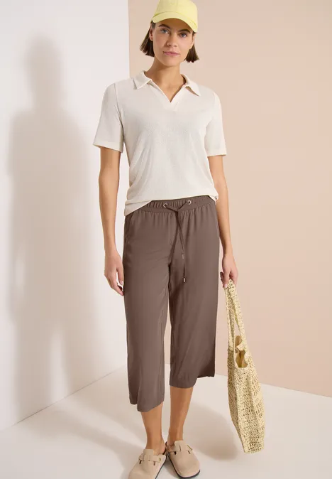 Vrouw in wit poloshirt en bruine 7/8-broek met gevlochten boodschappentas, casual look.