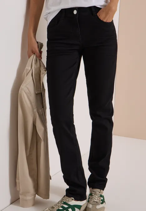 Jean noir droit; modèle tient un manteau beige et porte des baskets à motif
