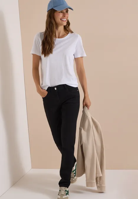 Femme en t-shirt blanc, jean noir et casquette bleue, tenant une veste beige devant un fond neutre.