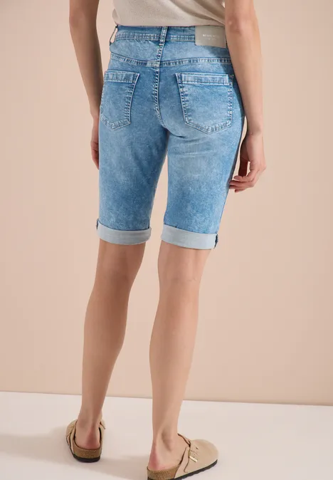 Vrouw van achteren in lichtblauwe, knielange jeansshorts met omslag en beige loafers tegen een lichte achtergrond.