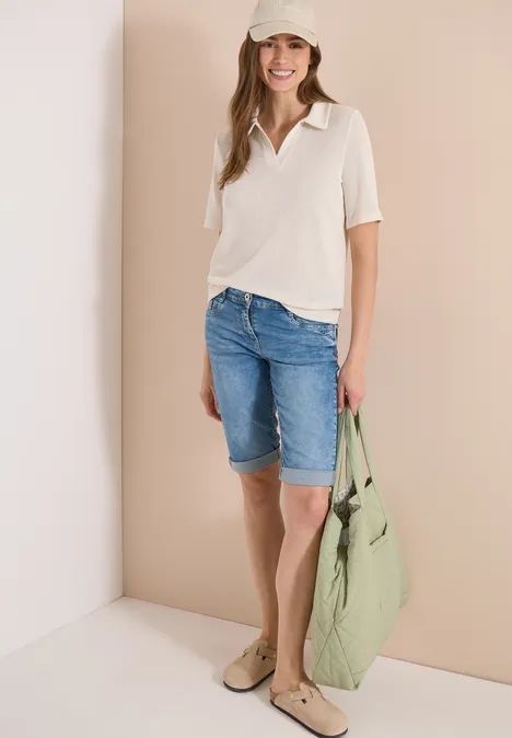 Vrouw in licht shirt, jeansshorts en grote groene tas tegen een beige muur.