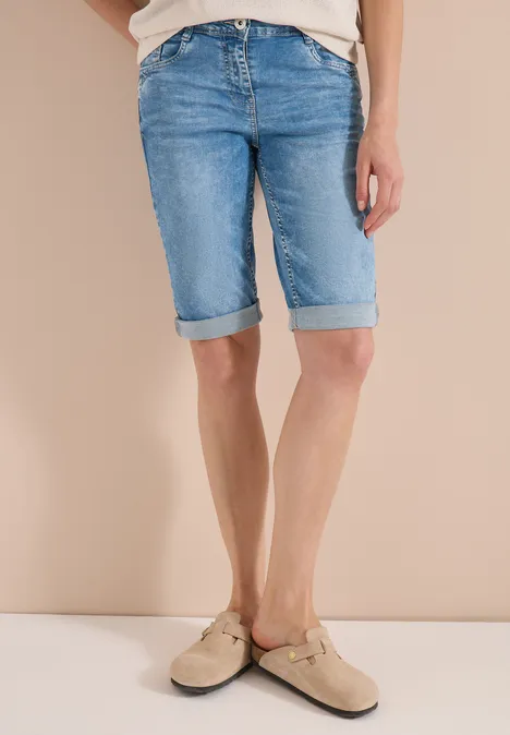 Persoon draagt lichtblauwe, omgeslagen denim bermudashort en beige instappers voor een beige achtergrond.