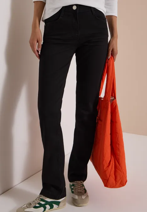 Pantalon noir évasé, baskets vertes et blanches, doudoune orange à la main.