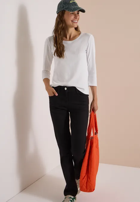 Femme souriante en t-shirt blanc et jean sombre, sac rouge à la main, appuyée contre un mur