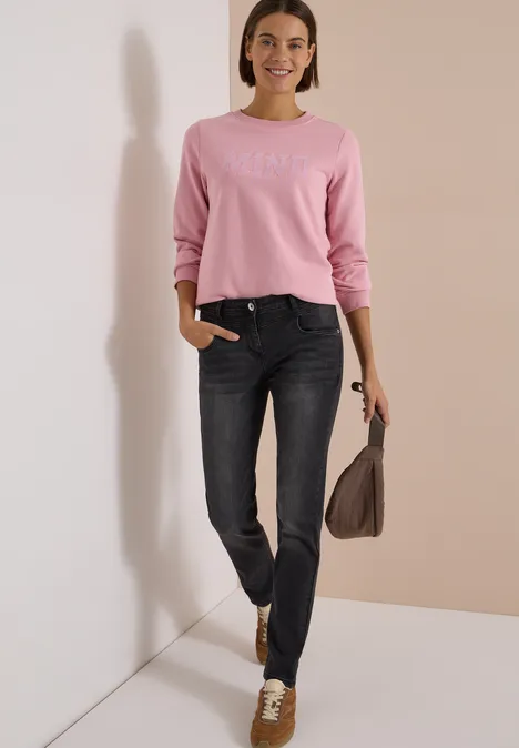 Eine Frau in rosa Pullover und dunklen Jeans steht lächelnd vor einer Wand und hält eine braune Tasche.