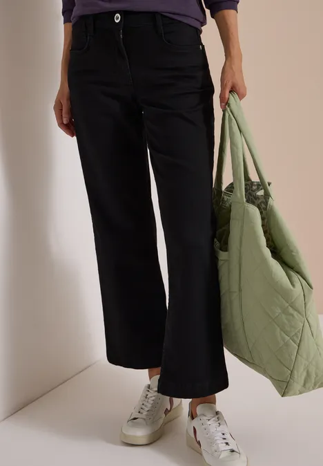 Vue des jambes en jean noir et baskets blanches, avec un sac matelassé vert.