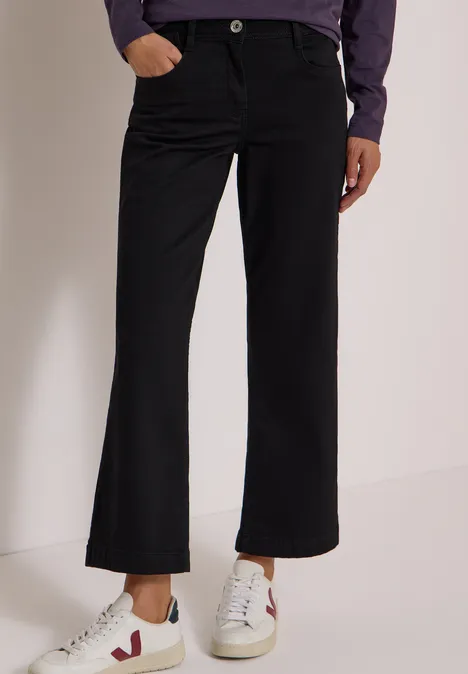Pantalon noir ample pour femme avec fermeture à bouton, porté avec baskets blanches sur fond neutre.