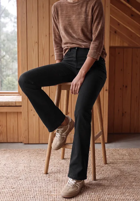 High Waist Bootcut Jeans im Slim Fit Black Eine Frau sitzt entspannt auf einem Holzschemel, trägt schwarze Jeans, braunen Pullover und helle Sneaker.