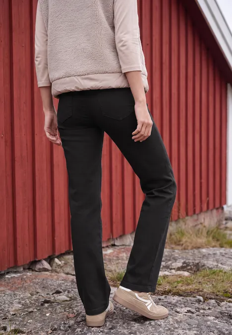 High Waist Wide Leg Jeans im Slim Fit Black Rückansicht einer Person in schwarzer Hose und beigen Schuhen vor einer roten Holzwand.