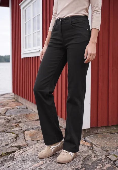 High Waist Wide Leg Jeans im Slim Fit Black Frau trägt schwarze gerade Jeans und beige Schuhe vor rotem Haus am See.
