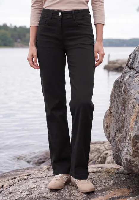 High Waist Wide Leg Jeans im Slim Fit Black Eine Person steht am Ufer auf Felsen, trägt schwarze, gerade geschnittene Hose und beige Schuhe.