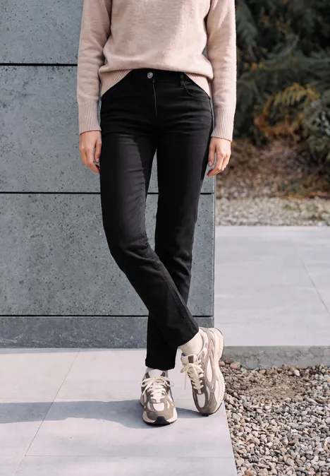 Person steht vor grauer Wand, trägt schwarze Hose und beige Sneakers, mit überschlagenen Beinen.