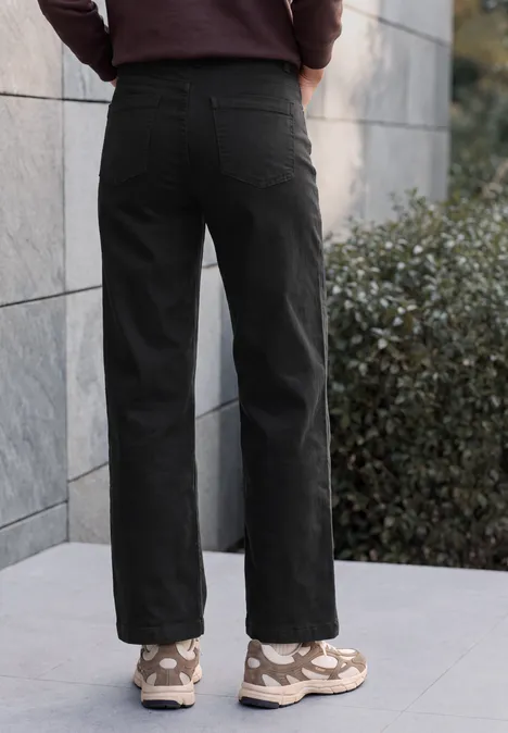 High Waist Wide Leg Jeans im Loose Fit Black Person von hinten in weiten schwarzen Hosen und beigen Sneakers vor grauer Steinwand.