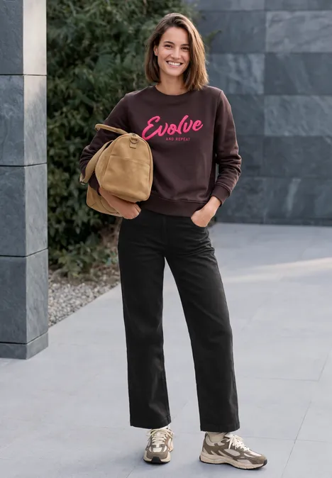 High Waist Wide Leg Jeans im Loose Fit Black Frau in schwarzer Hose und Sweatshirt ’Evolve’ steht lächelnd draußen mit Jacke in der Hand.