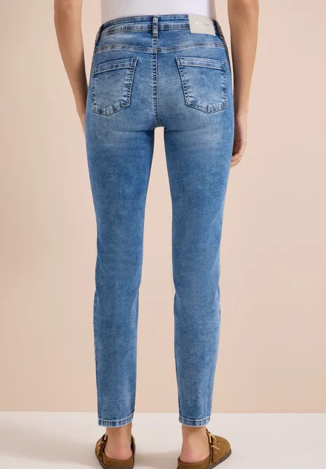 Rückansicht einer Person in hautengen blauen Jeans und Sandalen vor beigem Hintergrund.