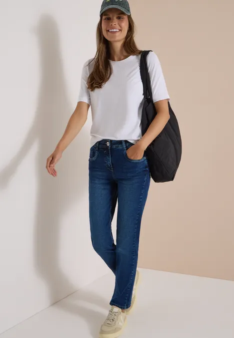 Jeans met High Waist en smalle pijpen in Slim Fit mid blue wash Glimlachende vrouw in wit T-shirt, blauwe jeans, groene pet en zwarte tas voor een lichte muur.
