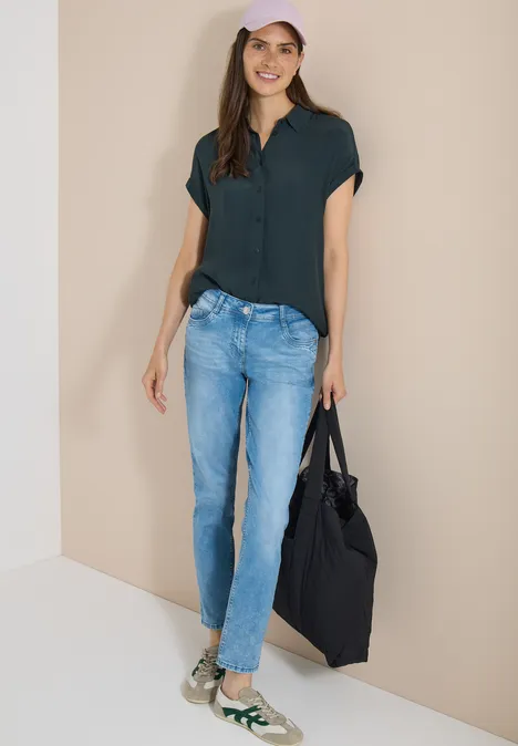 Jean taille moyenne, coupe Slim Leg et style décontracté light blue wash Femme en chemisier vert foncé, jean bleu, baskets vertes et sac noir sur fond beige.