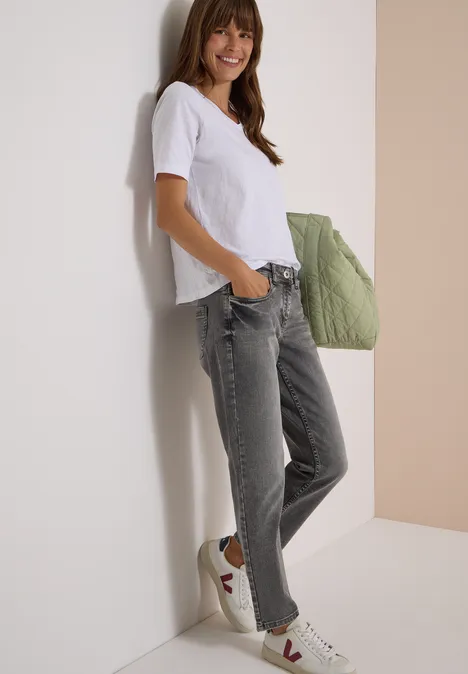 Jeans met Mid Waist-taille en Slim Leg-pijpen in casual pasvorm light grey wash Jonge vrouw leunt nonchalant tegen een muur, draagt wit t-shirt, grijze jeans, sneakers en een groene gewatteerde schoudertas.