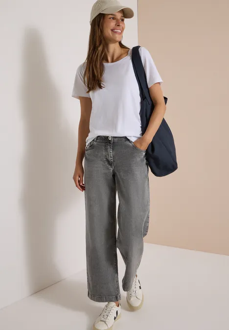 Jeans met High Waist en Wide Leg pijpen in een Loose Fit pasvorm light grey wash Vrouw in wit T-shirt, losse grijze jeans, sneakers, pet en schoudertas voor een neutrale achtergrond.