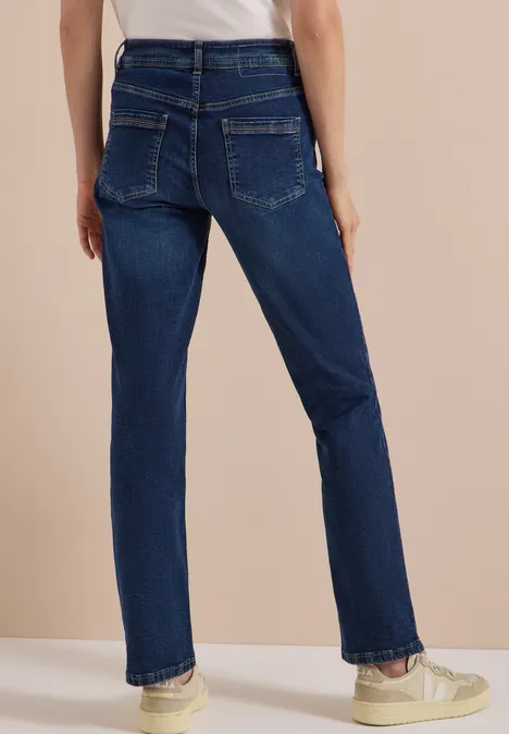 Rückenansicht einer Person in dunkelblauen, geraden Jeans und weißen Sneakers vor beige gehaltenem Hintergrund.
