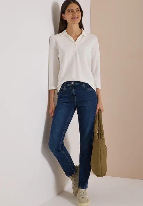 Frau lehnt lächelnd an Wand, trägt weiße Bluse, blaue Jeans und hält eine olivgrüne Tasche.
