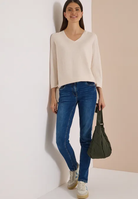 Frau in cremefarbenem Pullover, blauen Jeans und goldenen Sneakern mit grünem Beutel vor hellem Hintergrund.