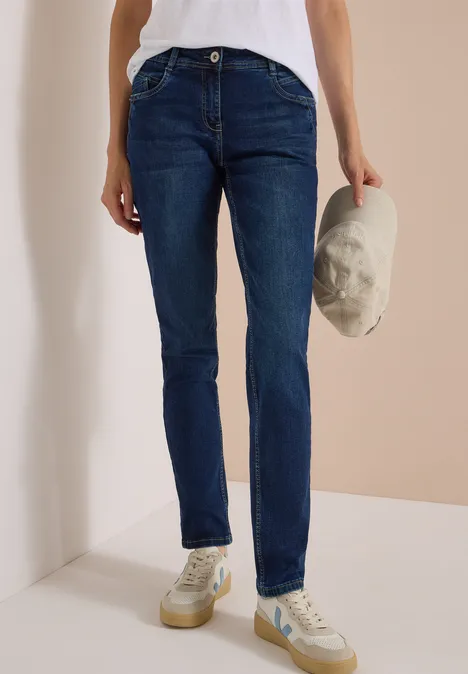 Person trägt dunkelblaue Jeans, weiße Sneakers und hält eine helle Kappe in der Hand.