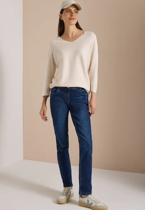 Eine Frau in dunkelblauen Jeans, beiger V-Pullover und Cap steht vor neutralem Hintergrund.