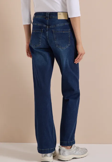 Rückenansicht einer Person in dunkelblauen, weiten Jeans und weißen Sneakers vor beigem Hintergrund.