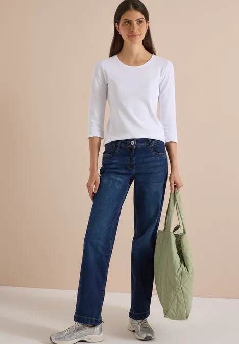 Eine Frau trägt ein weißes Langarmshirt mit 3/4-Ärmeln, weite Jeans und hält eine grüne Tasche.