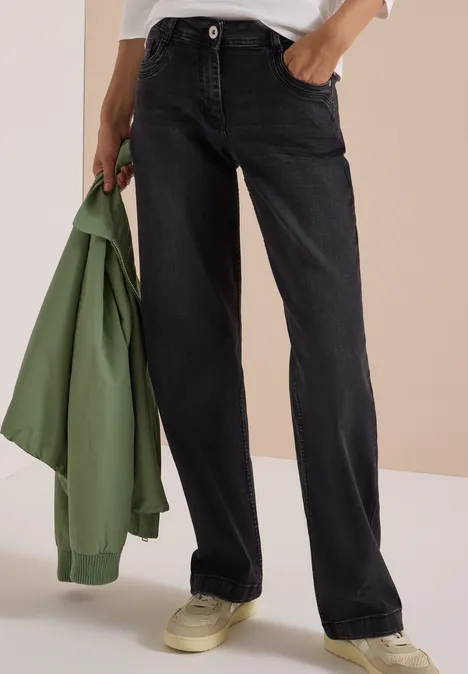 Person trägt schwarze, gerade geschnittene Jeans, hält in der linken Hand eine grüne Jacke und trägt helle Sneakers.
