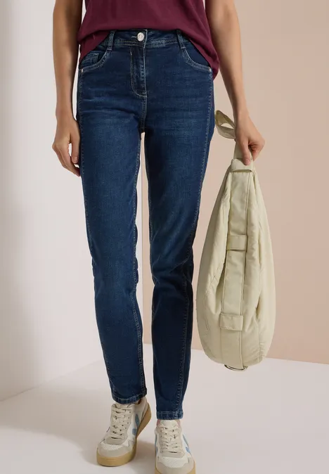 Femme en jean bleu foncé, baskets blanches, portant un sac beige à la main.