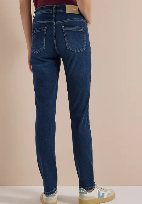 Personne de dos portant un jean slim bleu foncé et des baskets blanches devant un fond beige.